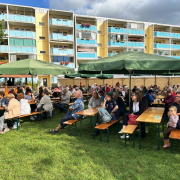 Menschen sitzen an Bierzeltgarnituren auf einer Wiese, im Hintergrund ist ein Wohnhaus zu sehen in denen Familien leben