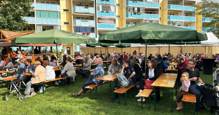 Menschen sitzen an Bierzeltgarnituren auf einer Wiese, im Hintergrund ist ein Wohnhaus zu sehen in denen Familien leben