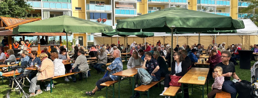 Menschen sitzen an Bierzeltgarnituren auf einer Wiese, im Hintergrund ist ein Wohnhaus zu sehen in denen Familien leben