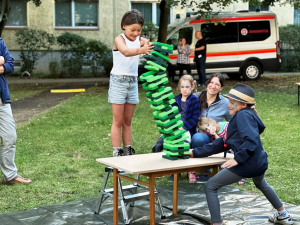 Kinder sind um einen kleinen Tisch auf einer Wiese und spielen Jenga mit grünen Holzbausteinen, der Turm ist gerade am umfallen