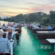 Mehrere Personen sitzen zusammen an unterschiedlichen Tischen auf einem Dampfer.