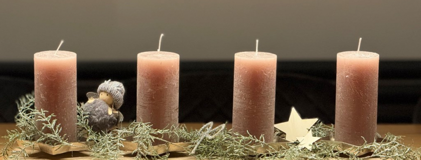 Auf einem Tisch steht längliches Adventsgesteck mit vier großen Kerzen. Darauf sind ein kleiner Engel, eine glitzerne Schleife und Sterne aus Holz dekoriert.