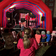 WG Solidarität Kinder sitzen auf Bänken vor einer Bühne. Auf der Bühne steht ein Mann mit Weihnachtsmannmütze vor einem Mikrofon.