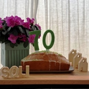 70 Jahre Geburtstag Ein Kuchen mit Kerzen in Form einer siebzig steht auf einem Tisch. Dahinter stehen Blumen sowie drei kleine Häuser aus Holz. Vor dem Kuchen ist das Wort SOLI zu sehen, welches aus Holzbuchstaben besteht.