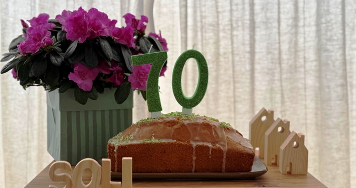 Ein Kuchen mit Kerzen in Form einer siebzig steht auf einem Tisch. Dahinter stehen Blumen sowie drei kleine H&auml;user aus Holz. Vor dem Kuchen ist das Wort SOLI zu sehen, welches aus Holzbuchstaben besteht.