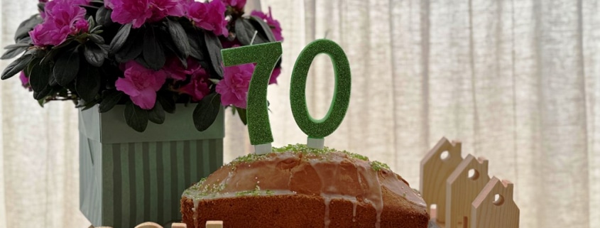 Ein Kuchen mit Kerzen in Form einer siebzig steht auf einem Tisch. Dahinter stehen Blumen sowie drei kleine Häuser aus Holz. Vor dem Kuchen ist das Wort SOLI zu sehen, welches aus Holzbuchstaben besteht.