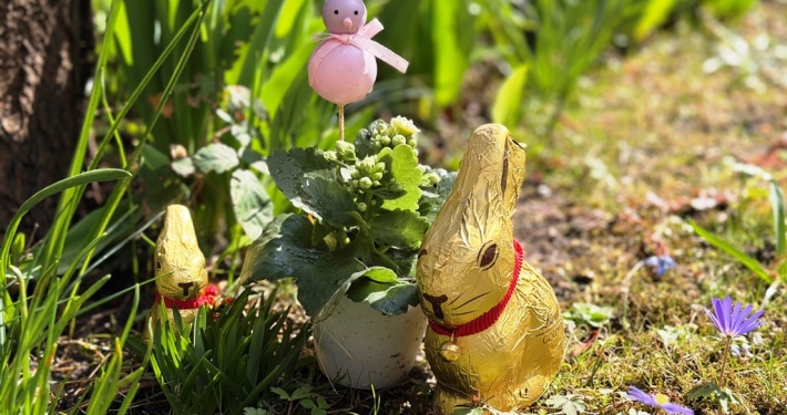 zwei goldenen Osterhasen sitzen auf einer Wiese. Ein Dekohase in rosa und lila ist zwischen Blumen zu sehen.