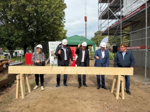 f&uuml;nf Personen stehen auf einer Baustelle hinter einem Holzbalken, vier davon schlagen mit dem Hammer einen Nagel in den Balken