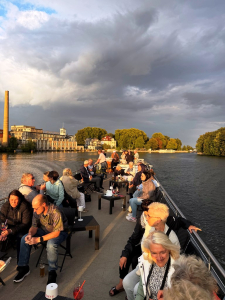 eine Gruppe von Menschen auf einem Dampfer. Im Hintergrund sind Geb&auml;ude, Wasser und viele B&auml;ume zu sehen.